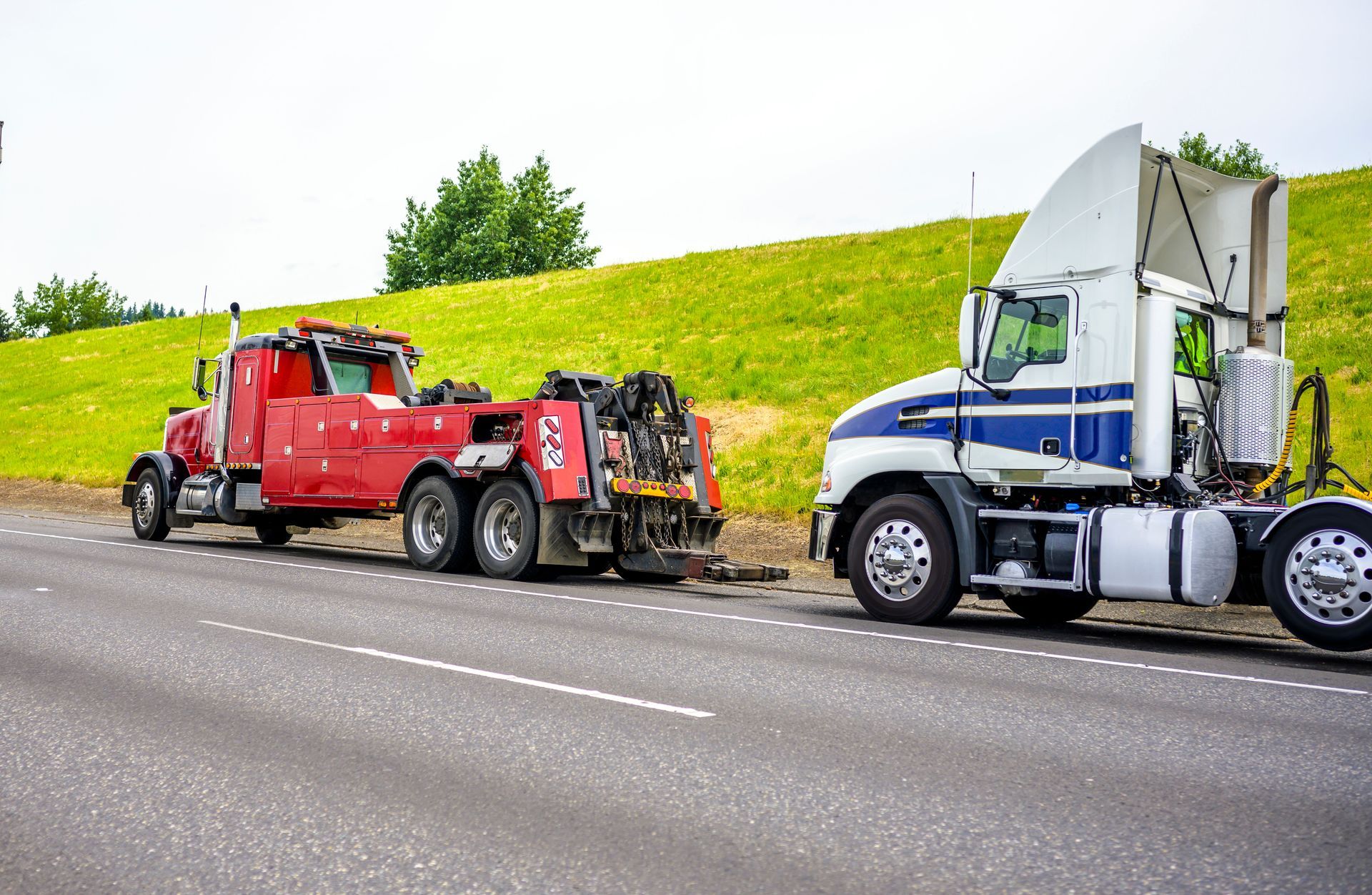 Red tow truck and blue semi truck parked on roadside beside grassy embankment