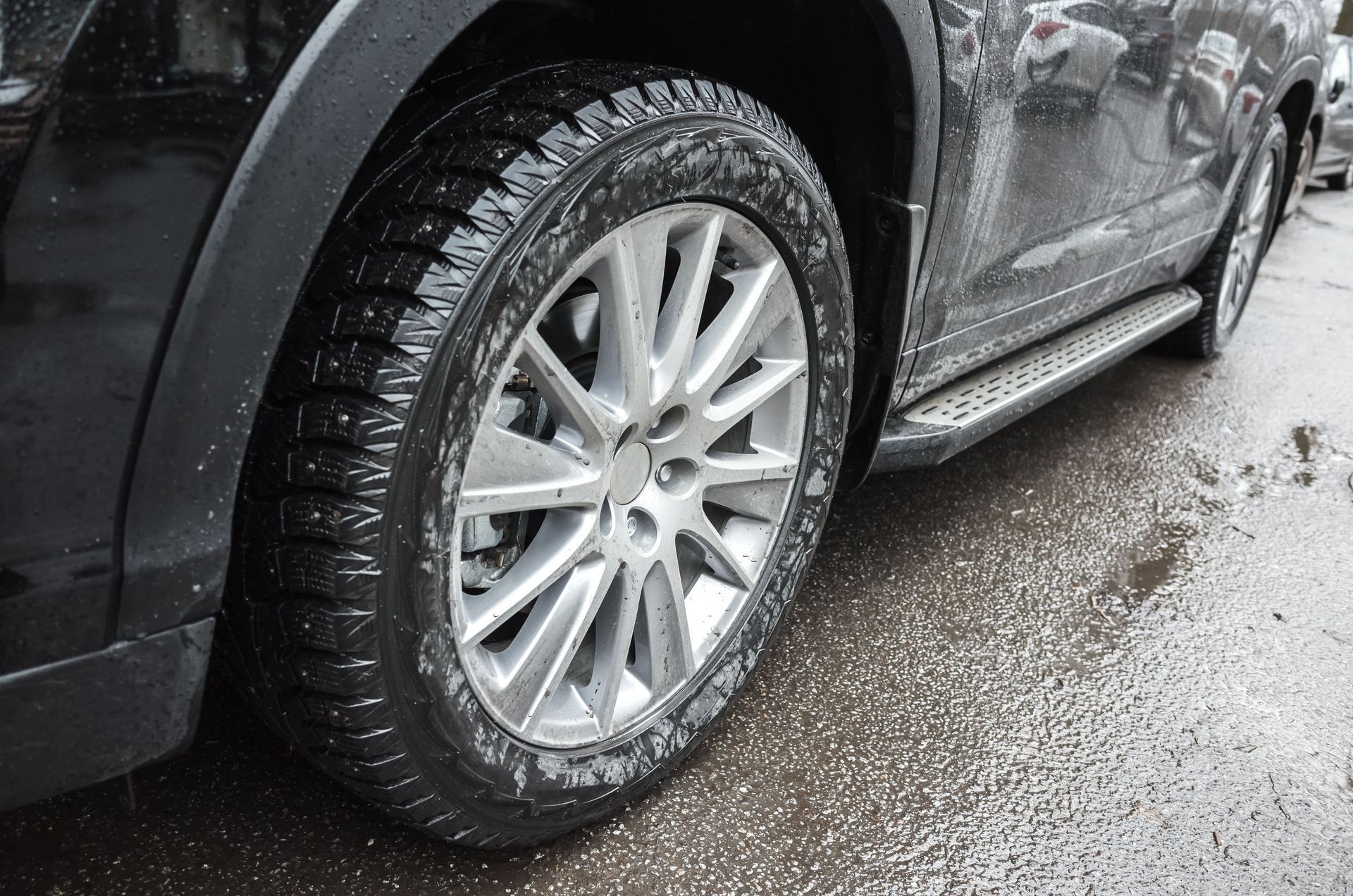 Close-up of a black car’s front wheel and silver rim on a wet road in rainy weather