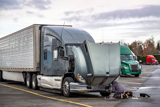 Semi truck with front damage in a parking lot, mechanic inspecting it on the ground.