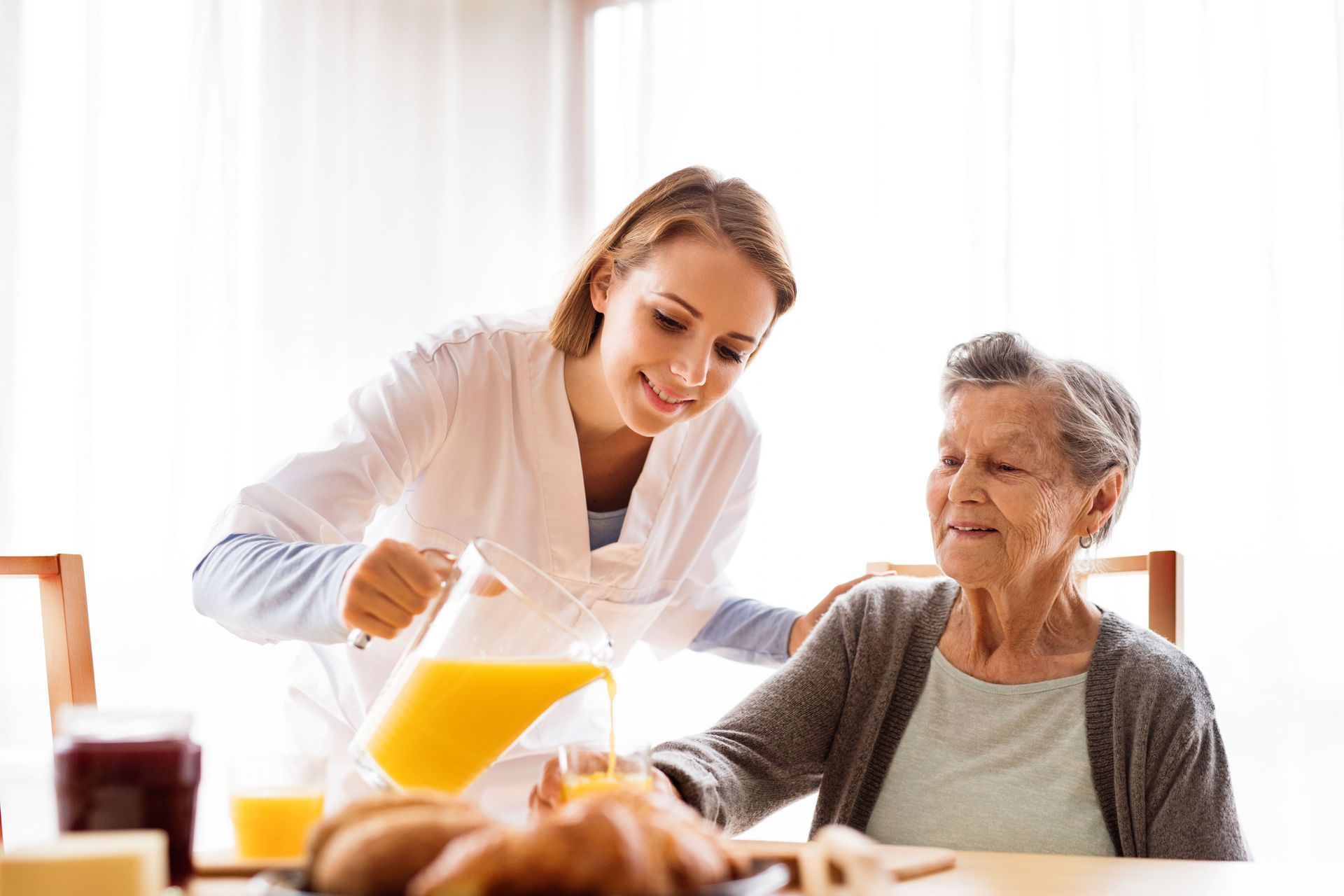 Caregiver pouring orange juice for an older woman at a dining table in a bright room