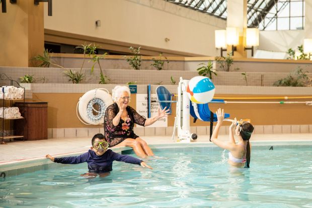 People in an indoor pool with a blue-and-white beach ball and a child floating on a kickboard