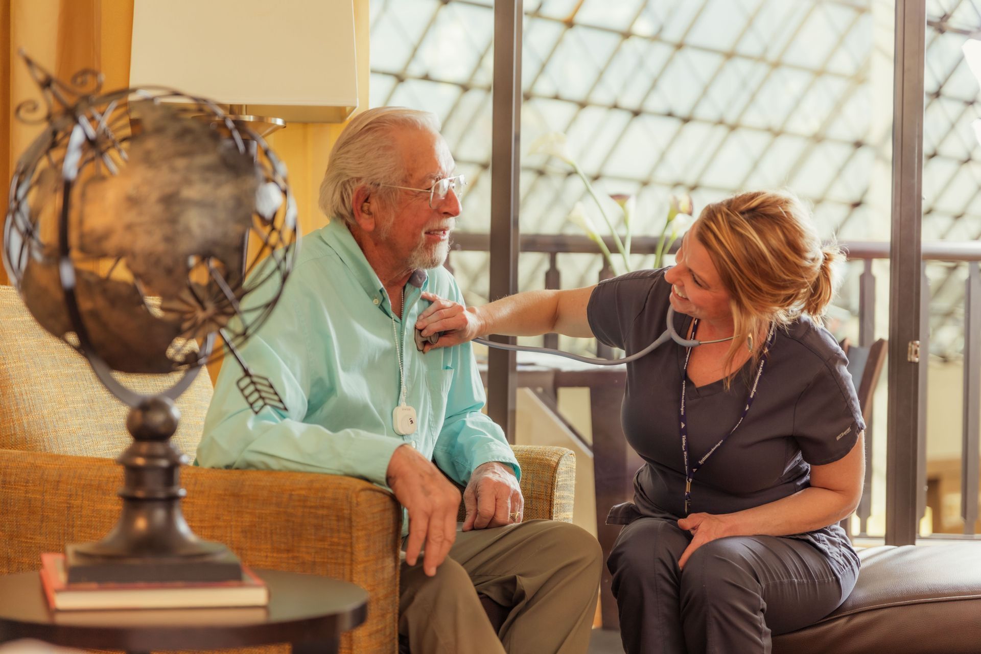 Caregiver checking an elderly man’s pulse on a couch in a bright living room