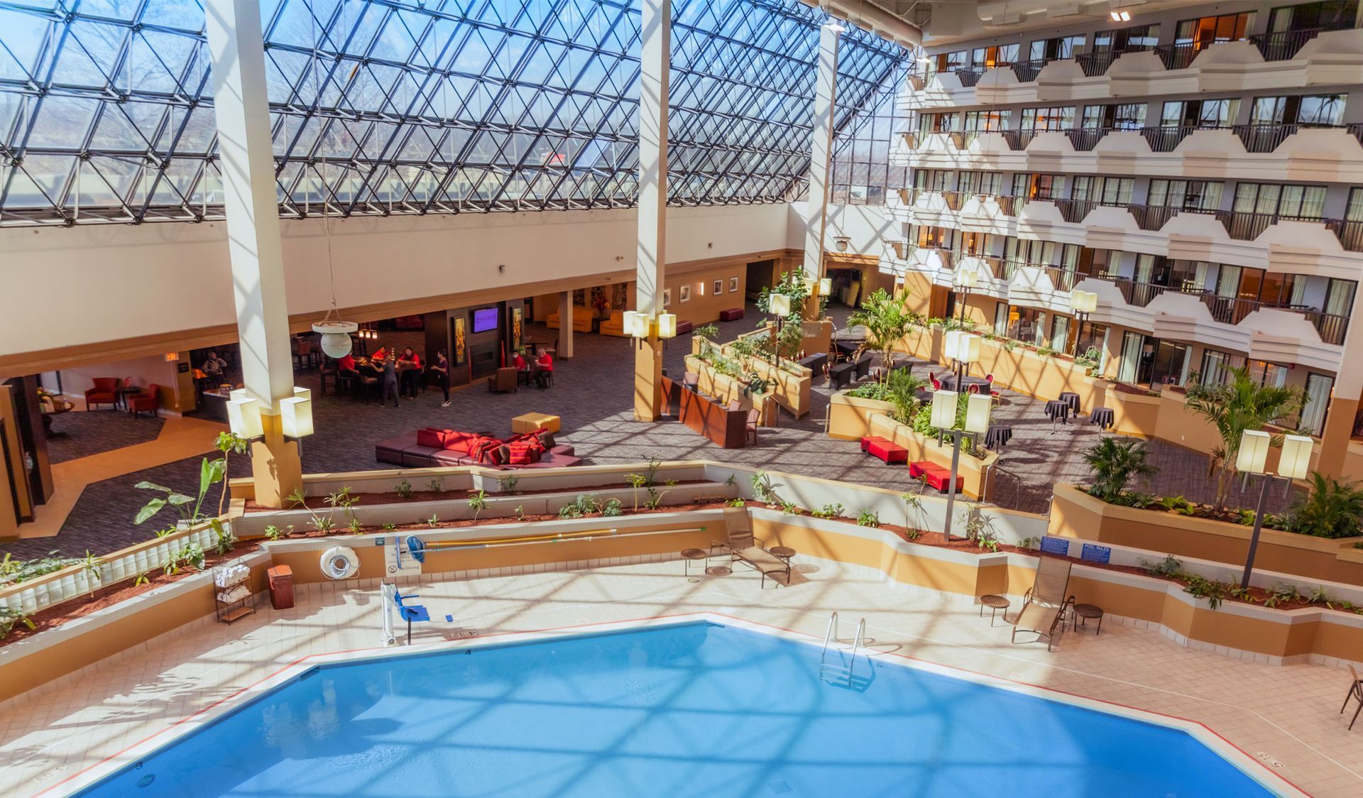 Indoor hotel atrium with a large blue pool, glass roof, balconies, and lounge seating