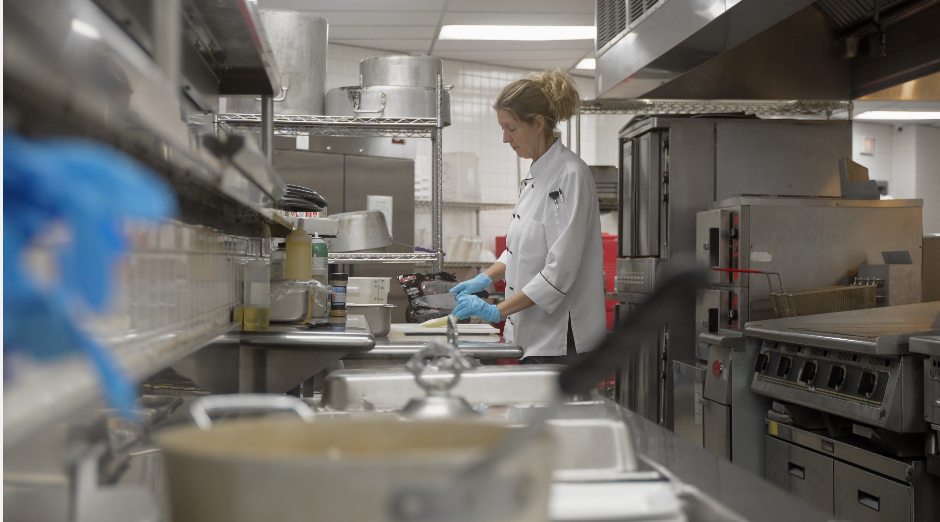 Chef preparing food in a stainless-steel commercial kitchen, with pots and utensils in the foreground.