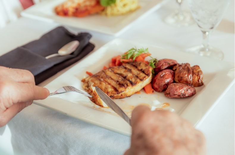Hands cutting grilled steak and sausage on a white plate at a formal dining table