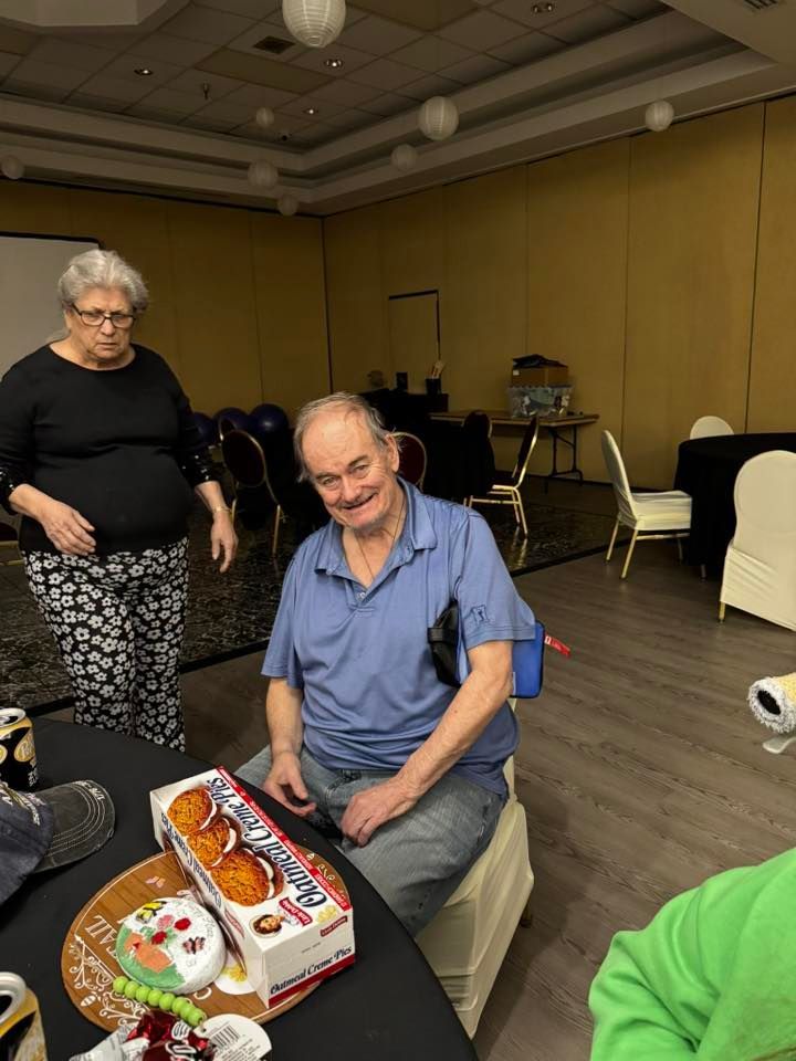 Man sitting by a table with a decorated cake while a woman stands nearby in a banquet room