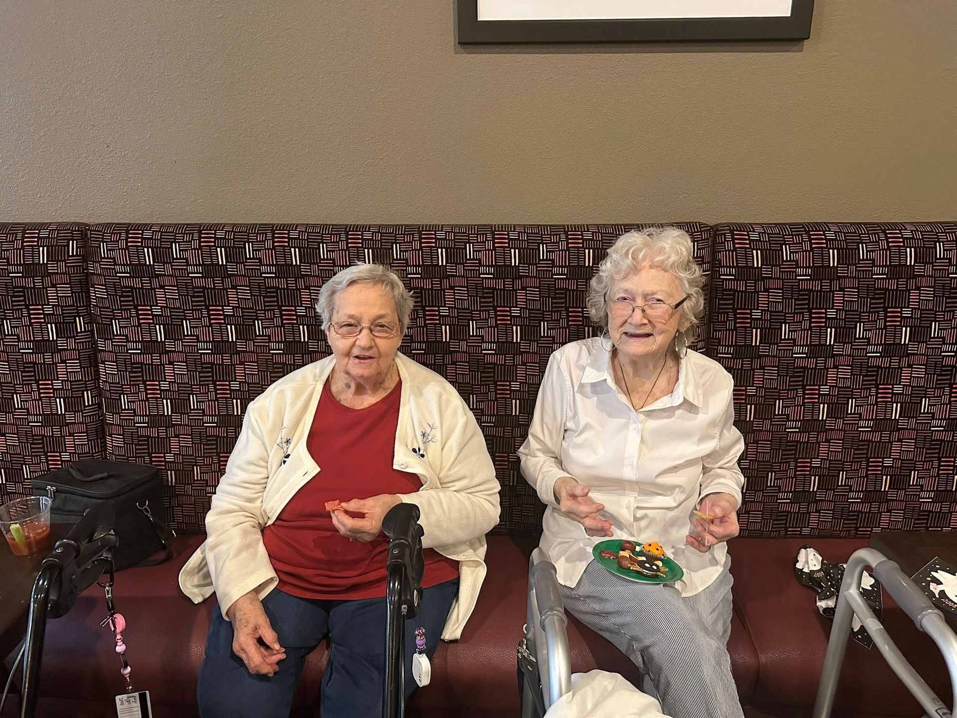 Two elderly women sit on leather chairs in a lounge, one holding a cane and a snack plate.