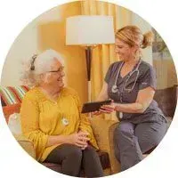 Nurse talking with an elderly patient in a yellow top beside a lamp in a warm living room.
