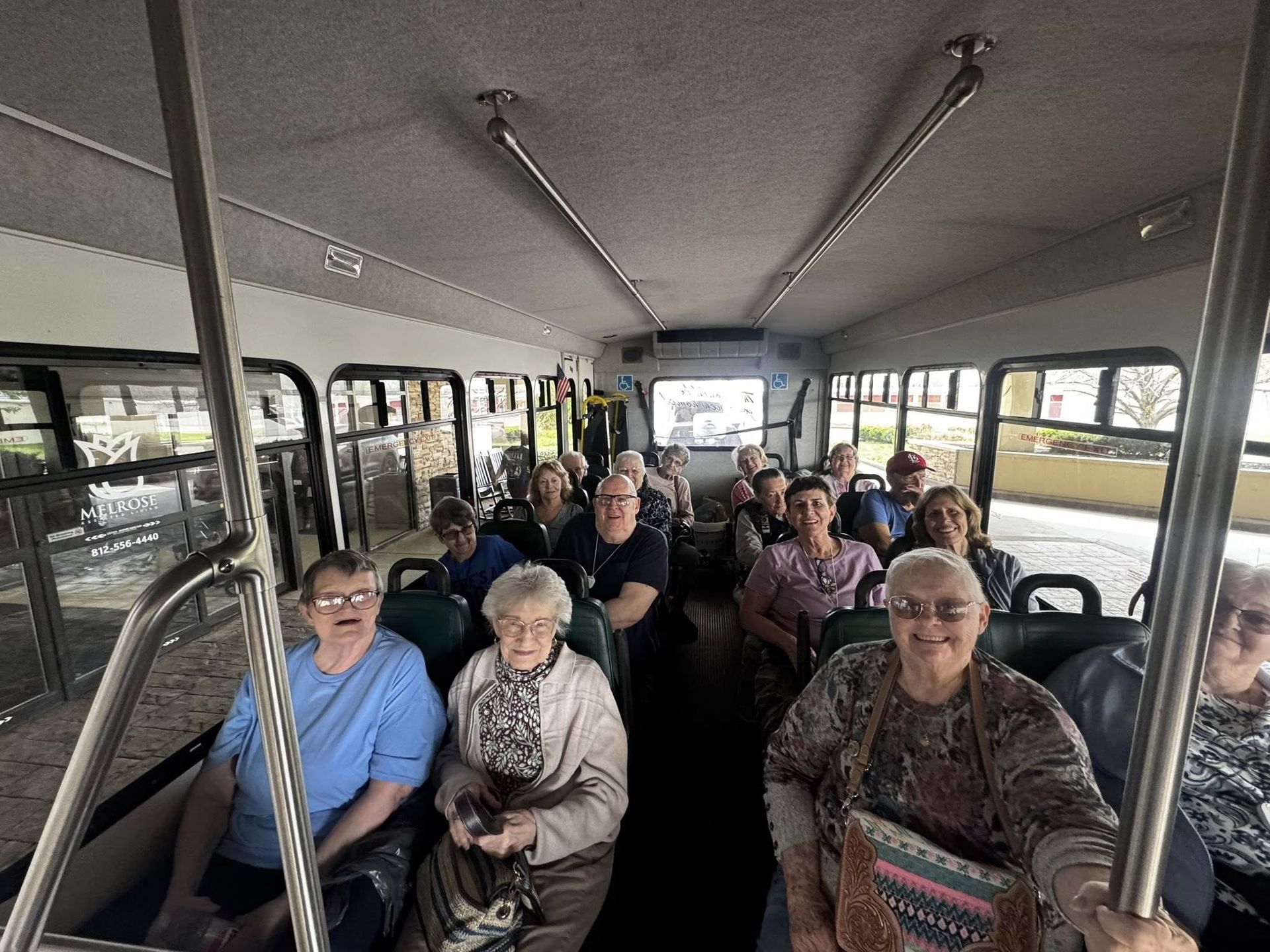 Group of people seated inside a tour bus, facing the camera with metal poles and windows visible.