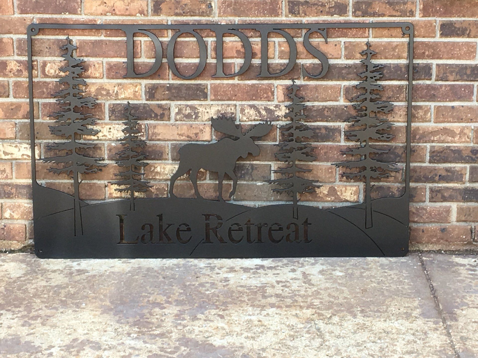 A metal sign with a moose and trees on it is hanging on a brick wall.