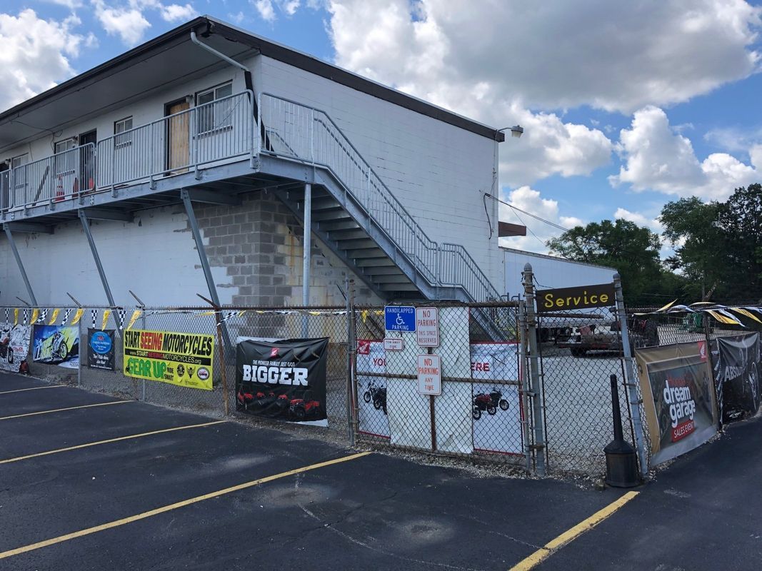 A white building with stairs leading up to the second floor is behind a chain link fence.