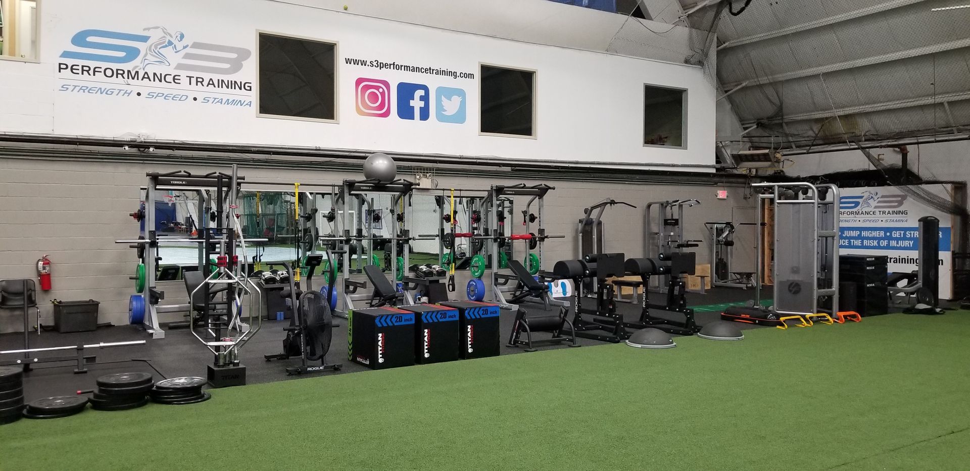 Gym with equipment, including weights, machines, and turf flooring. Social media logos are visible on the wall.