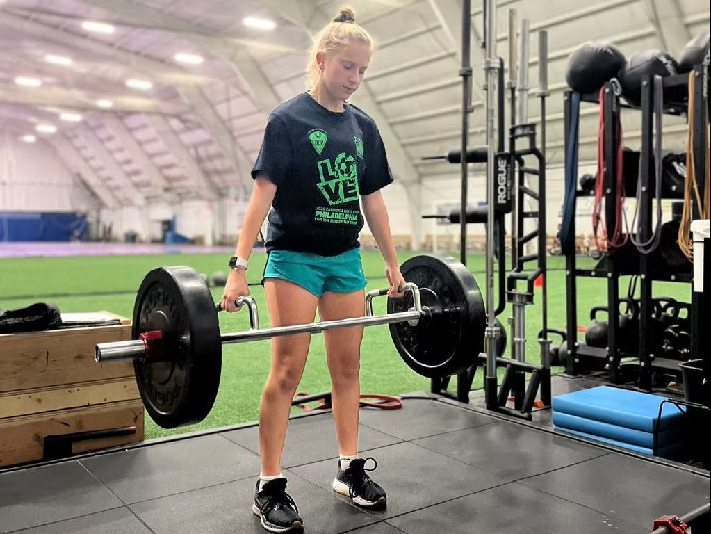 Woman deadlifting with a barbell in a gym. She wears a green shirt, blue shorts, and black shoes.