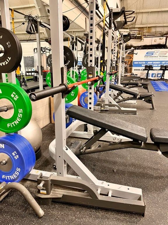 Weightlifting equipment in a gym, including a power rack, bench, barbells, and weight plates.