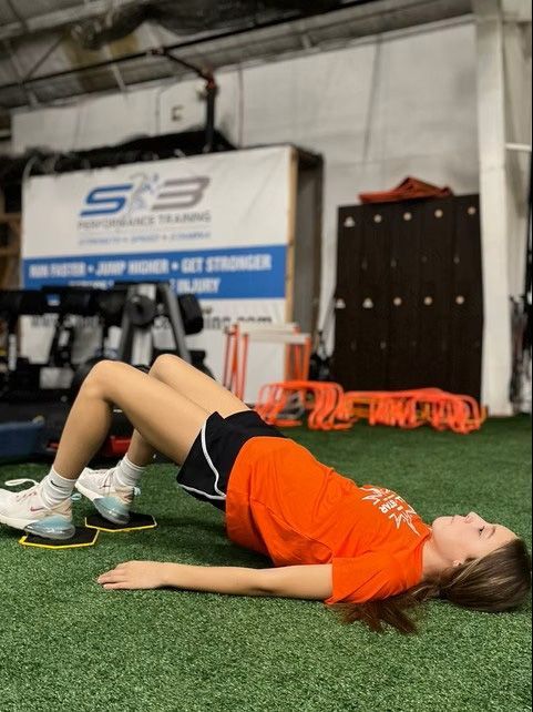 Woman doing a glute bridge exercise on a gym floor, with sliders under her feet. She wears orange top, black shorts.
