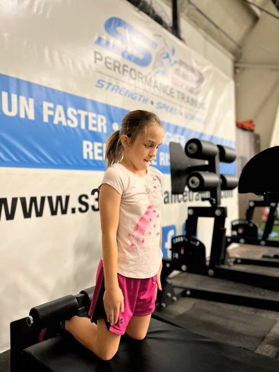 Girl in pink shorts kneels on a machine in a gym, looking down.