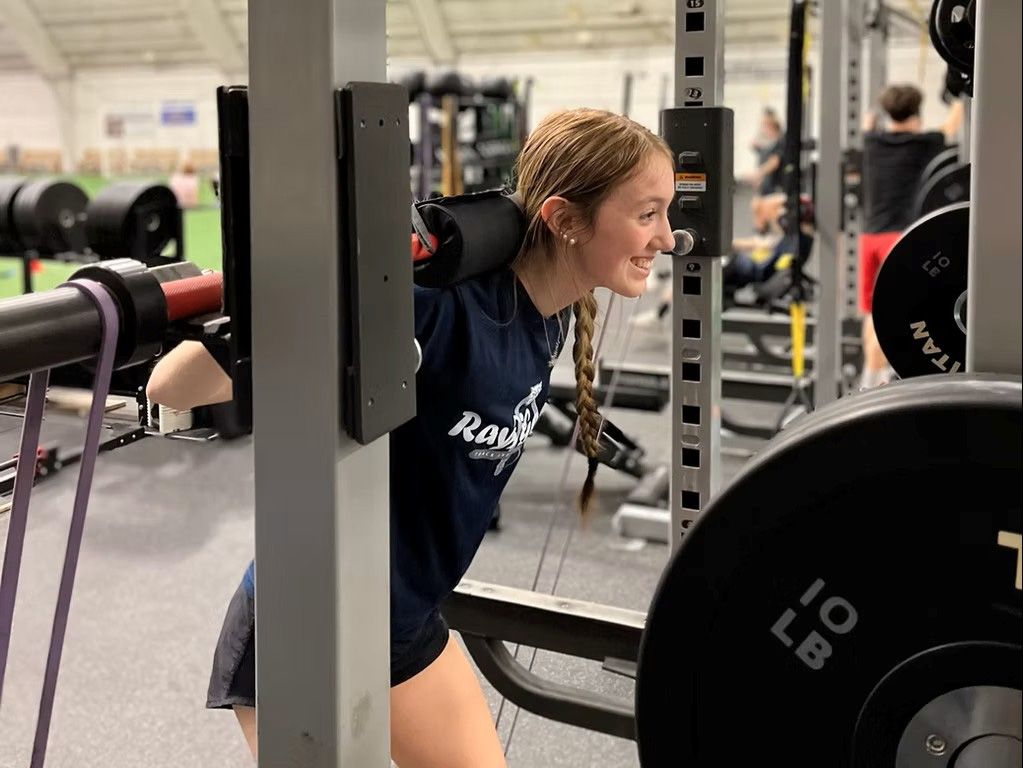 Woman with blonde braids squats in a gym, using a barbell and safety straps.