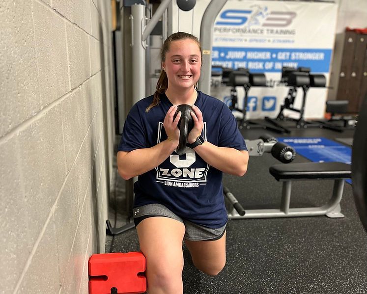 Woman in gym does a lunge, holding a weight. She wears a navy shirt and smiles, near a wall.