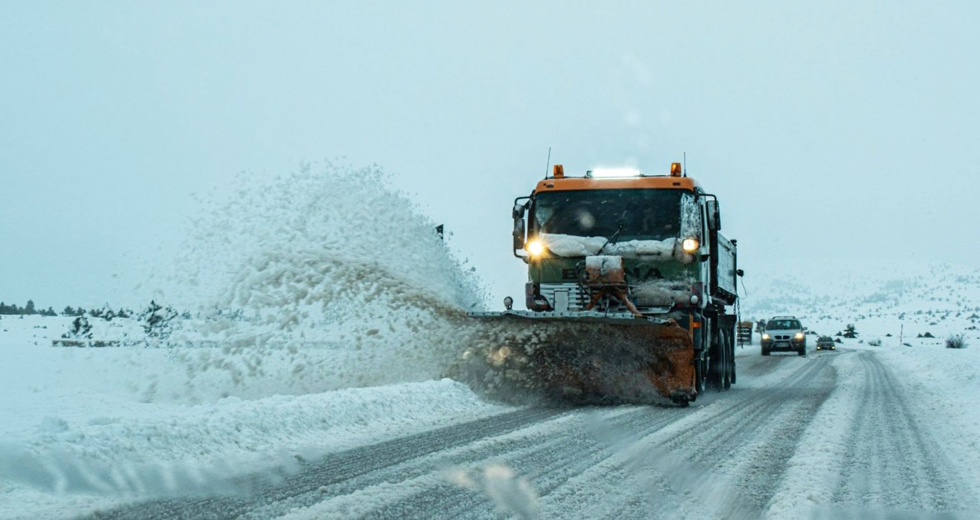 Snowplow clearing a snow-covered road; a car follows.