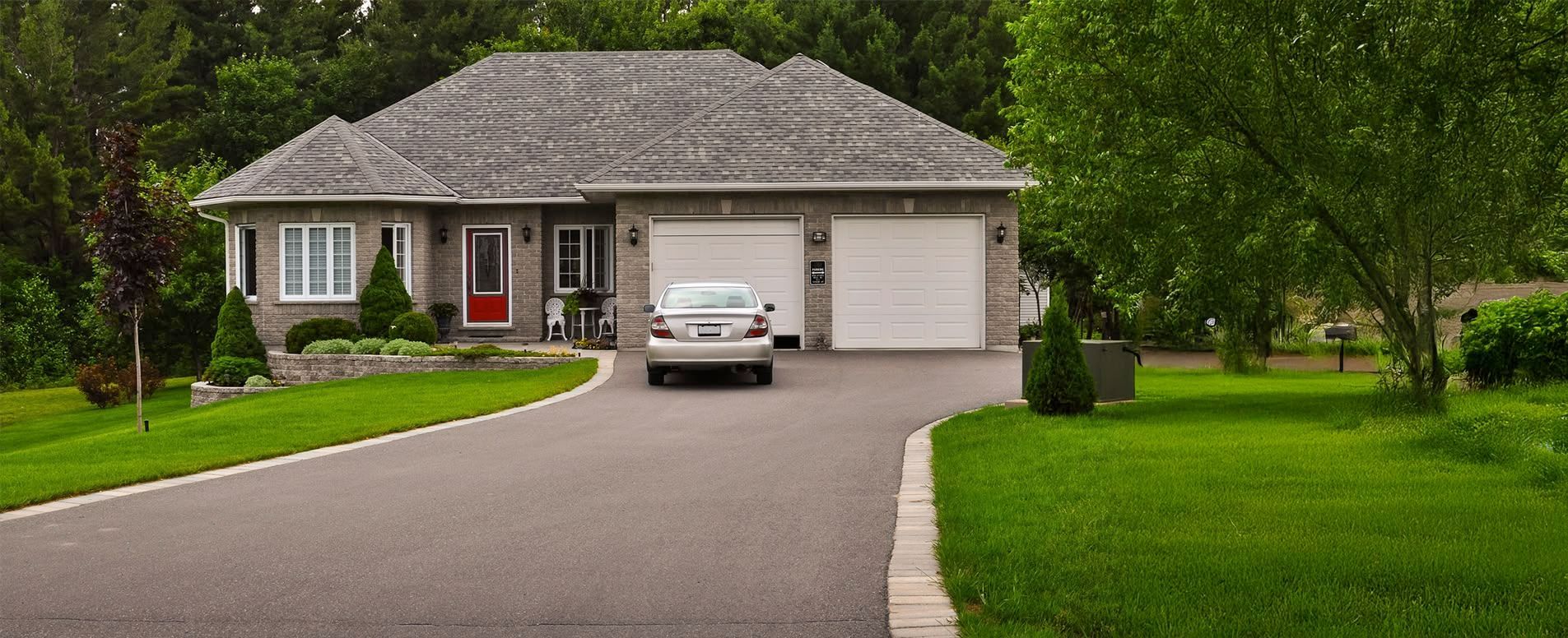 A gray house with a red front door, two garage doors, and a car in the driveway, surrounded by green grass and trees.