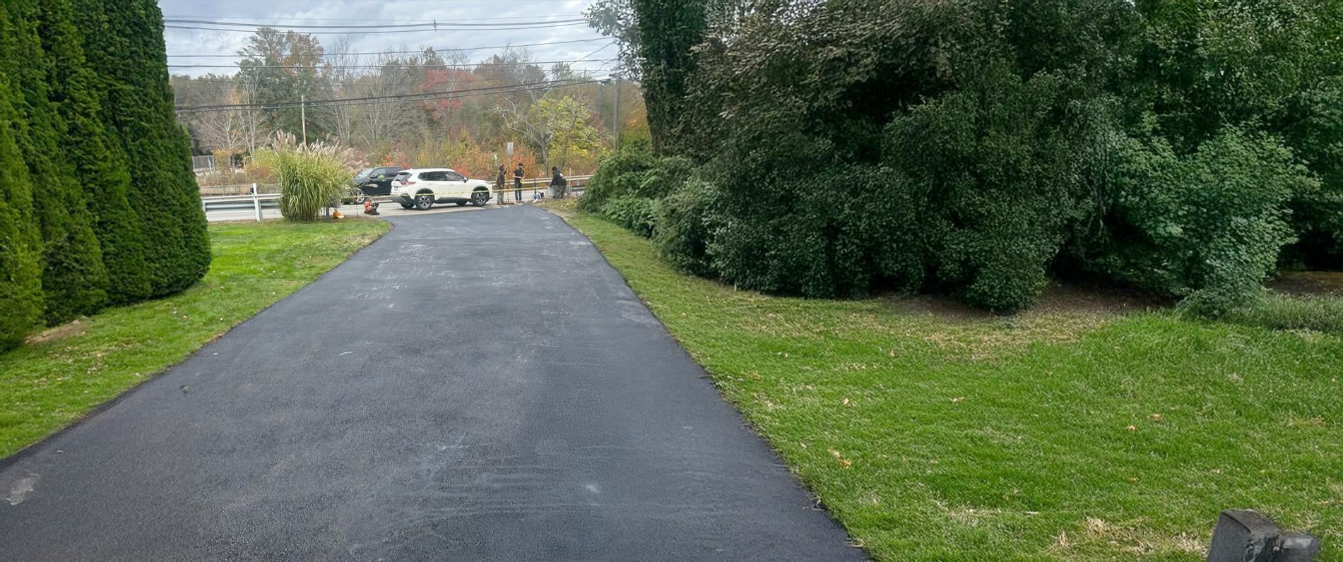 Asphalt driveway leads to a white truck parked near trees and lawn on a cloudy day.