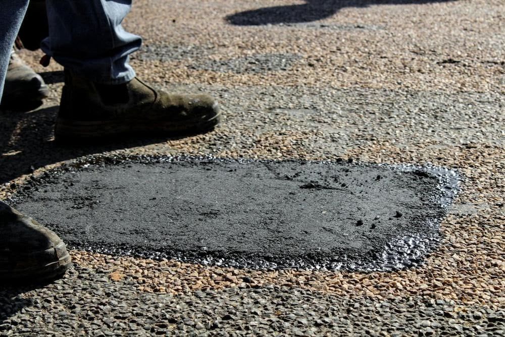 Patch of fresh asphalt on a road, near a person's feet.