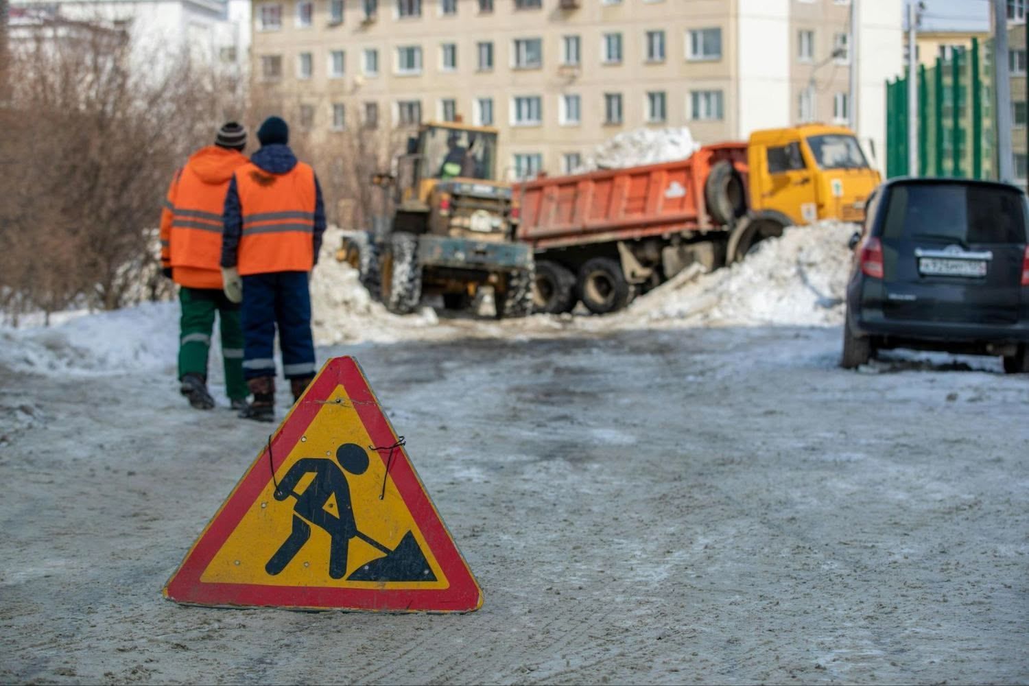 Construction sign on snowy road with workers, truck, and building in the background.