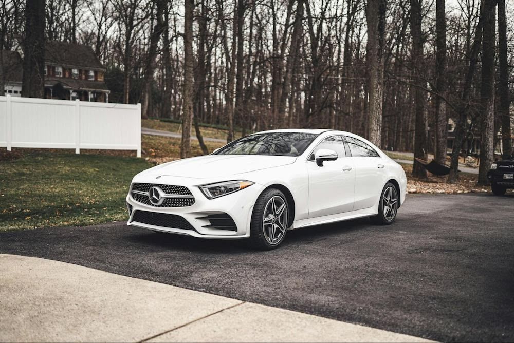 White Mercedes-Benz sedan parked on asphalt driveway, with bare trees and a house in the background.