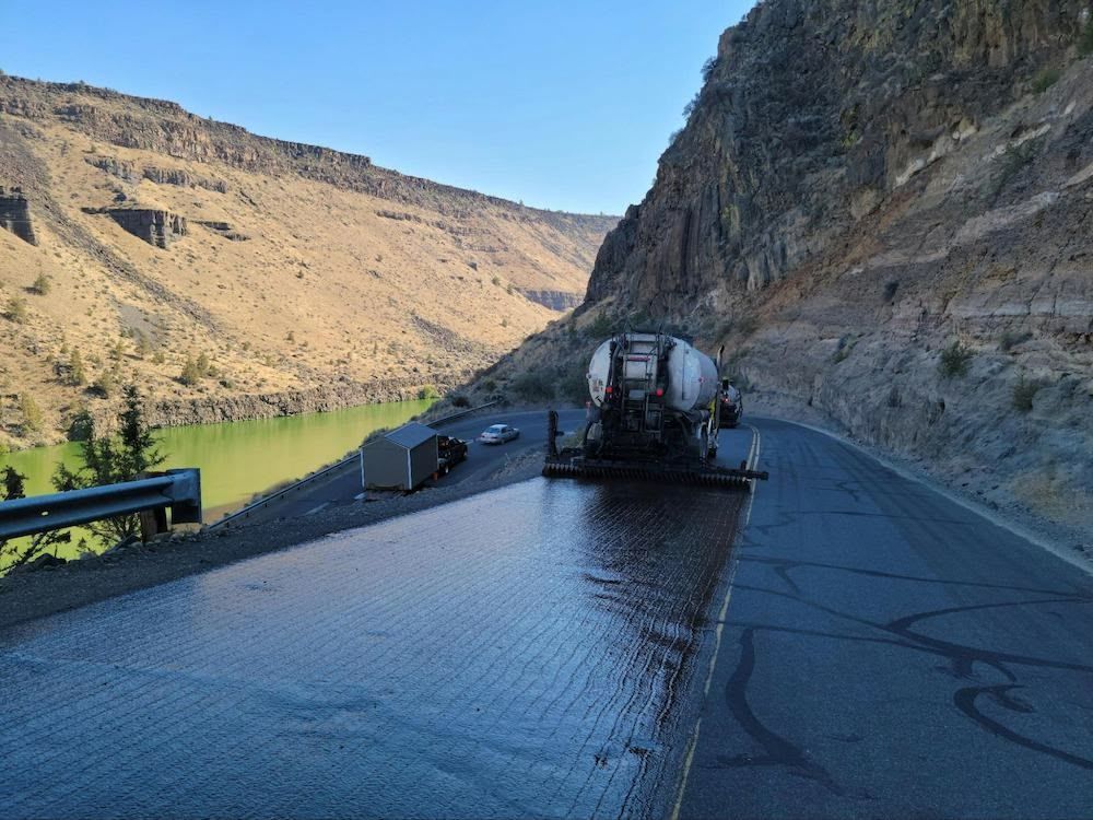 Road paving machine on a highway next to a river and cliffside.