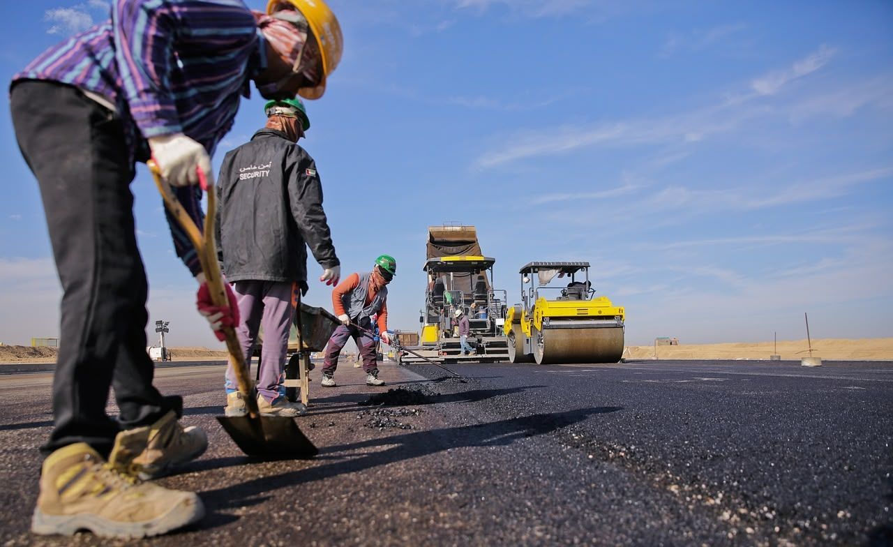 Construction workers paving a road on a sunny day. Two workers with shovels, machinery in background.