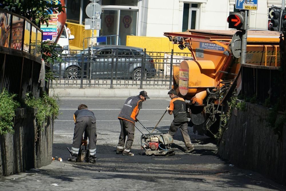 Road workers paving a street, with a machine, alongside a concrete wall.