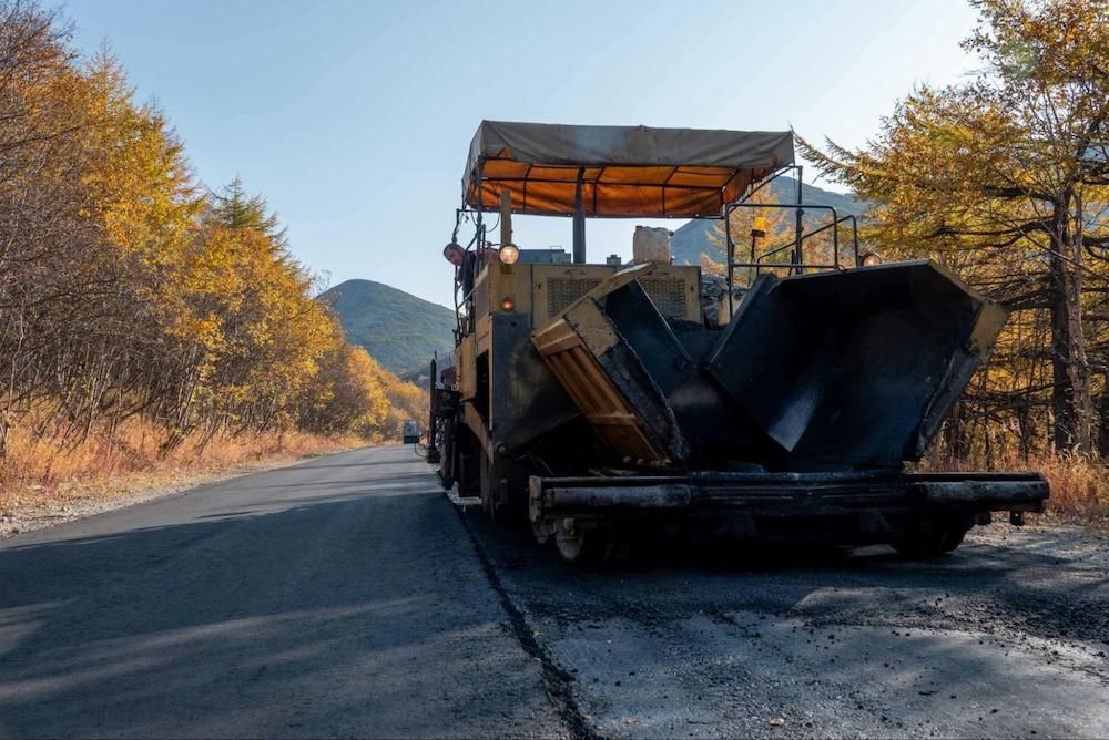 Asphalt paving machine on road, yellow and black, with trees in autumn colors.