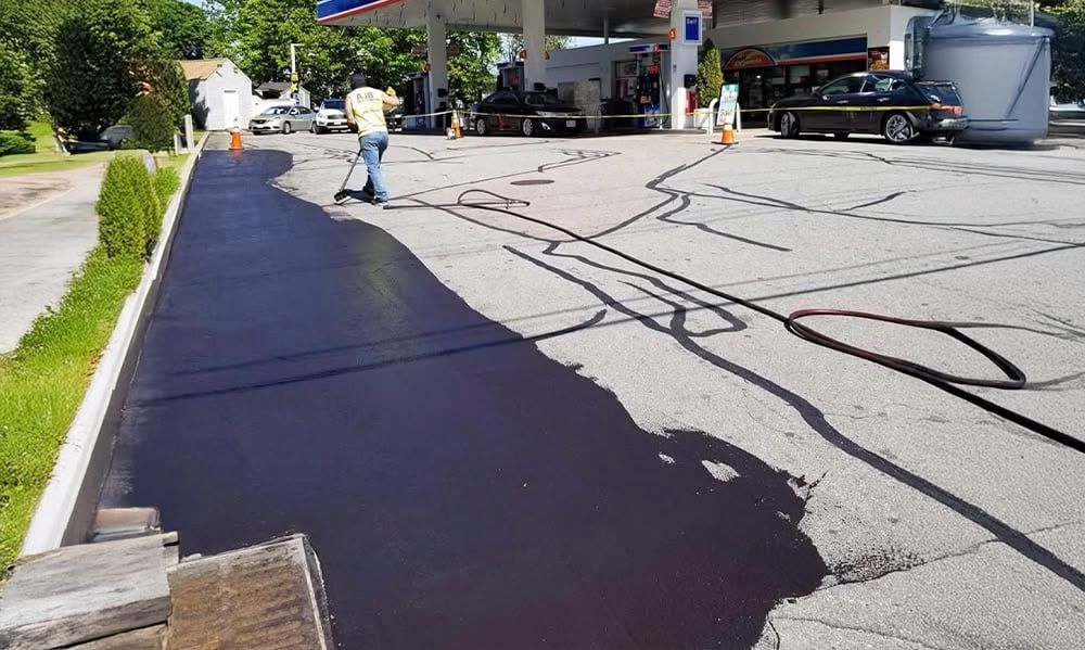 A person seals asphalt at a gas station. Black pavement and a worker with a squeegee are visible.
