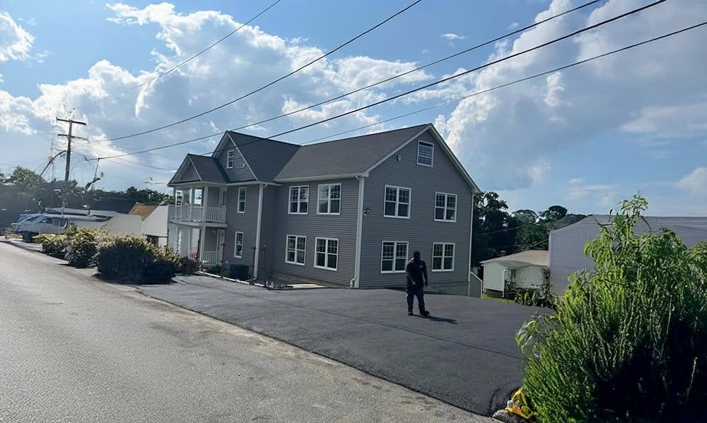 Gray two-story house with black asphalt driveway on a street with utility poles. Cloudy sky. Person standing.