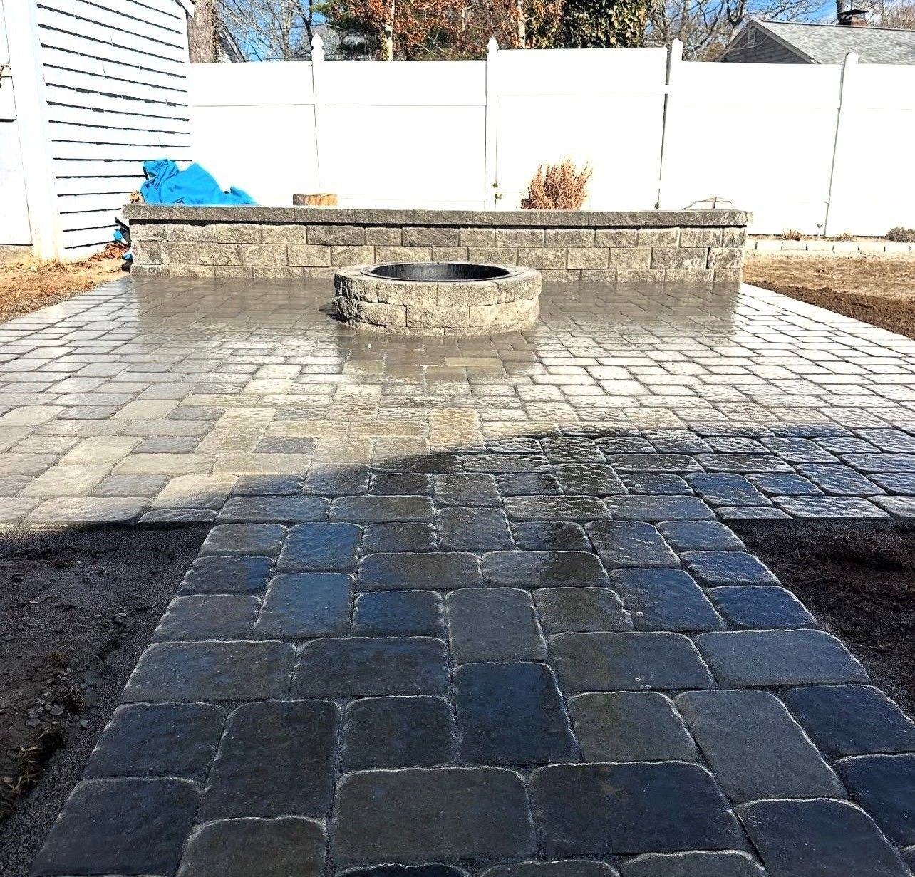 A stone patio with a central circular fire pit and a raised retaining wall in a backyard with a white fence.