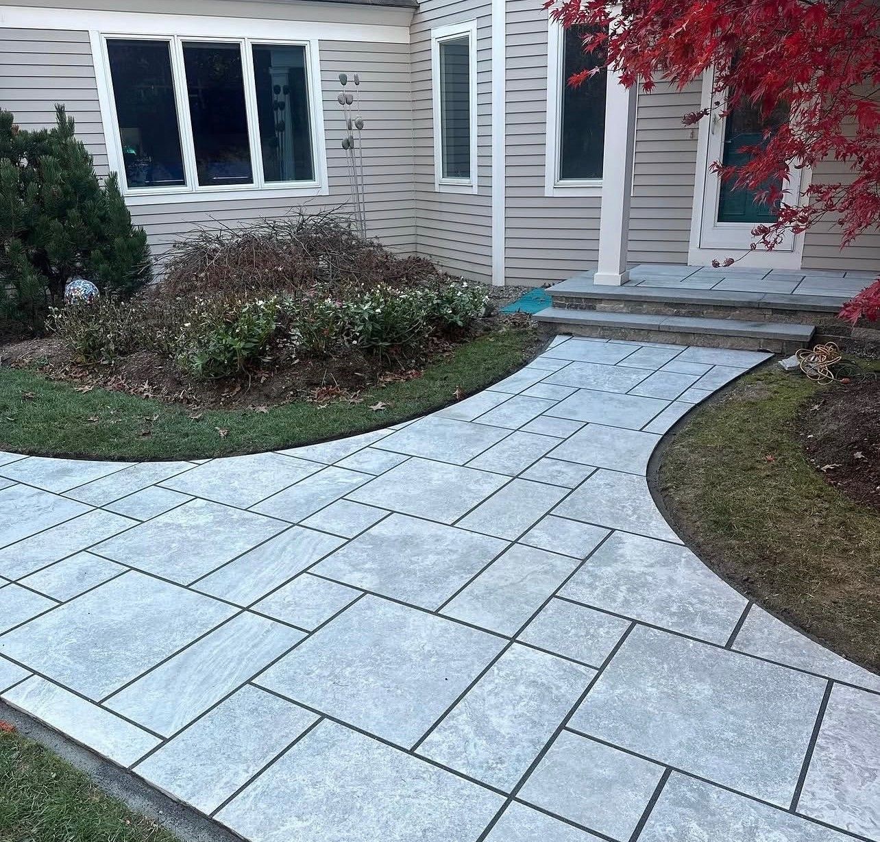 A stone walkway with a geometric pattern leading to a house entrance with light siding and red foliage.