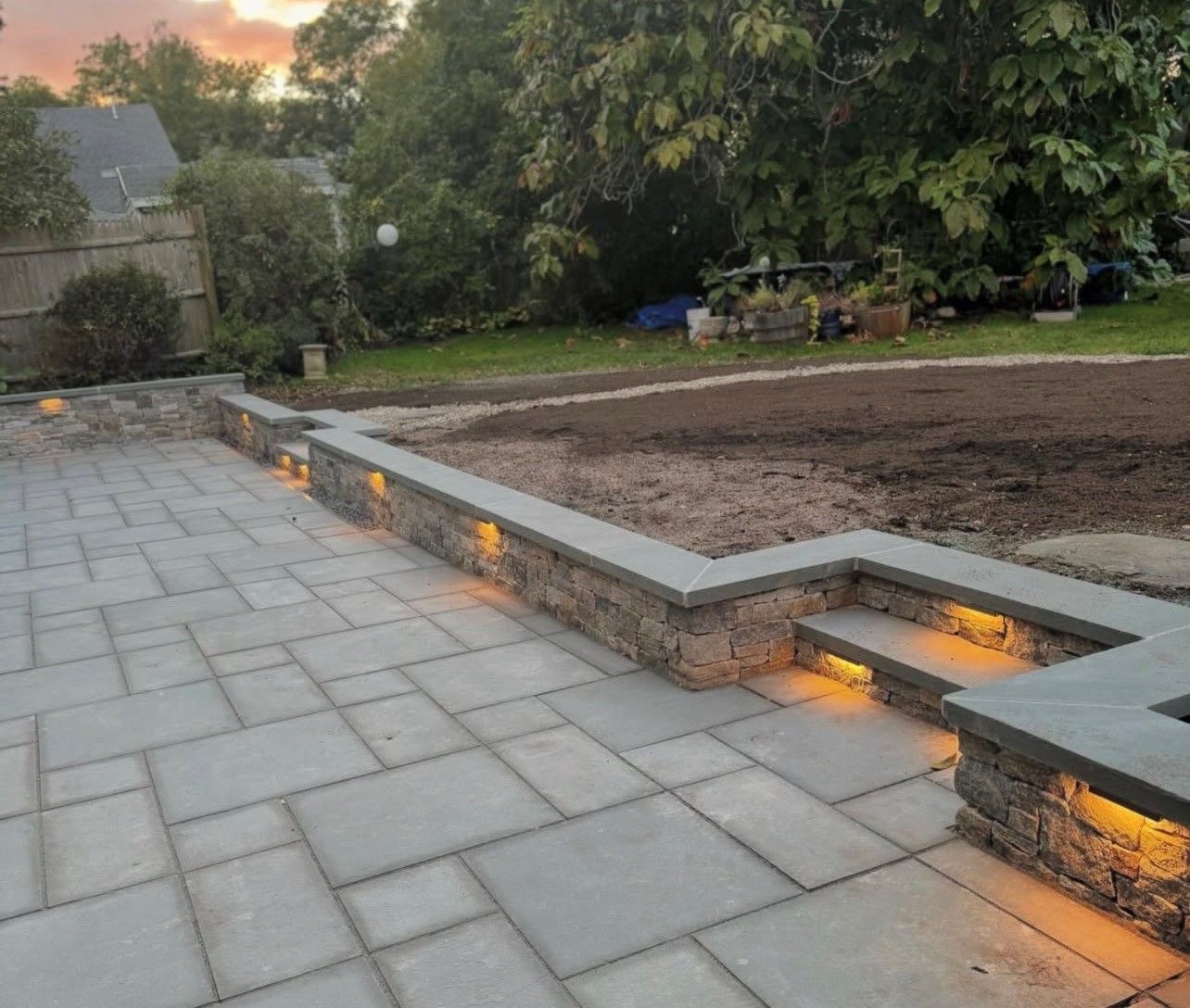 A stone patio with built-in retaining wall lighting at dusk, overlooking an unlandscaped yard with trees in the background.