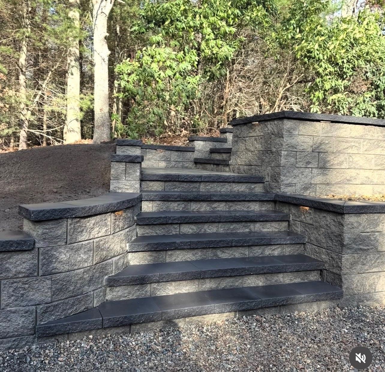 Stone steps with dark gray treads leading up a sloped landscape, flanked by matching retaining walls near a wooded area.