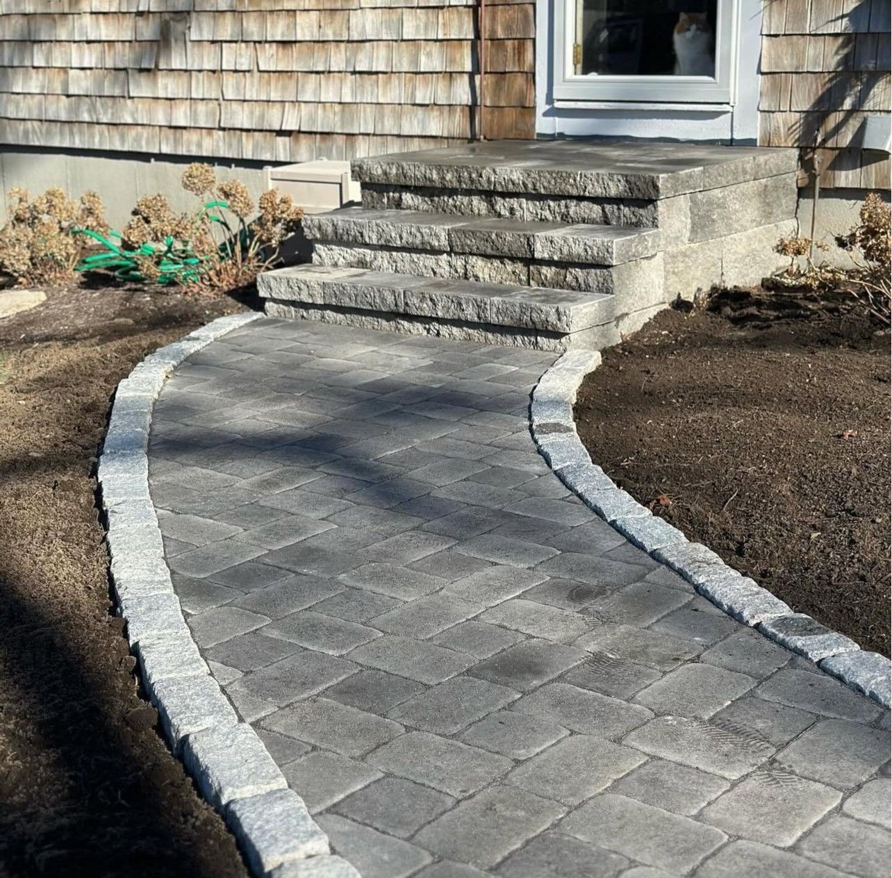 A curved stone paver walkway leads to a three-step gray stone staircase in front of a house with wood shingle siding.