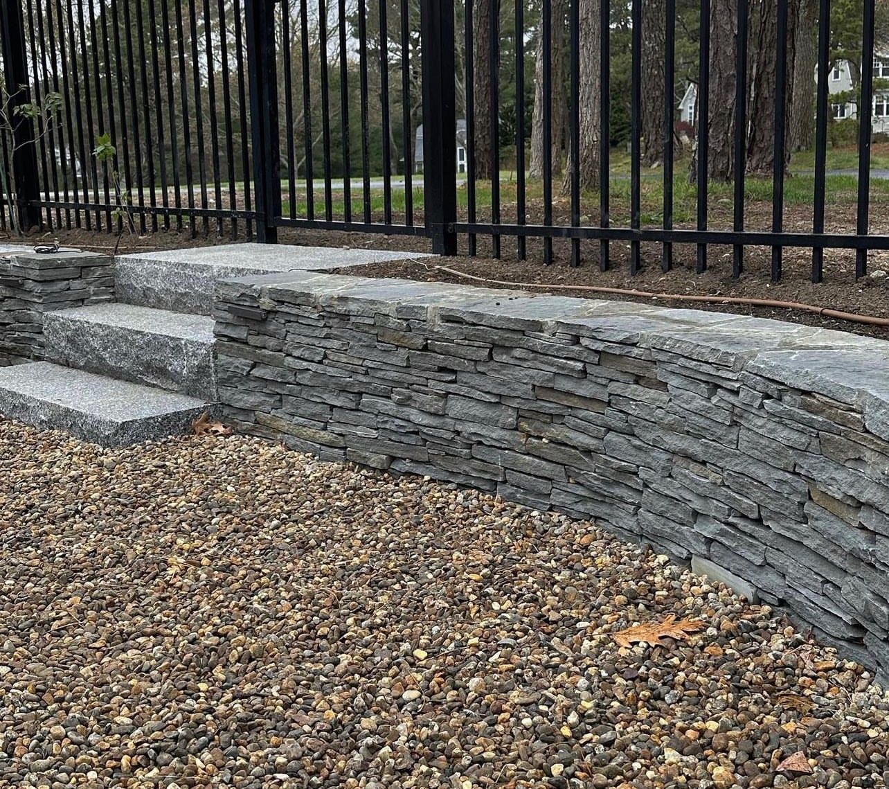 A curved, stacked stone retaining wall with integrated steps, set against a black metal fence and gravel ground.