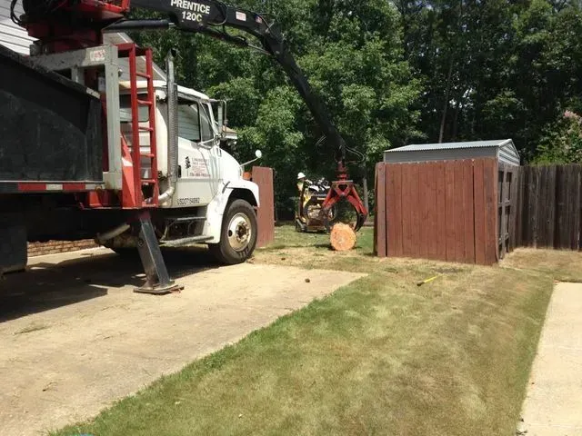 Truck with crane removing shed in a backyard with grass and a driveway.
