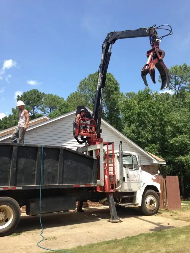 A truck with a crane and claw is removing debris from a house. A worker stands on the truck.
