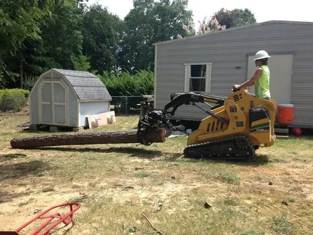 Person operating a small, yellow construction vehicle with a log in its jaws in a yard.
