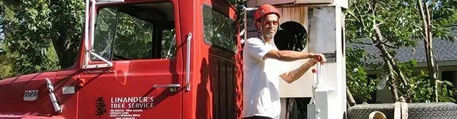 A red truck and two people working outdoors. One wears a hat and white shirt, gesturing. Green trees and a building are in the background.