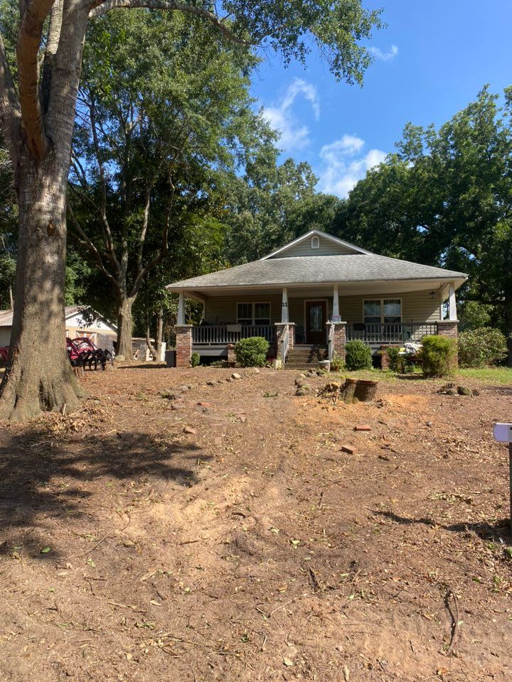 A one-story house with a porch. Brown and green landscape with a large tree on the left.