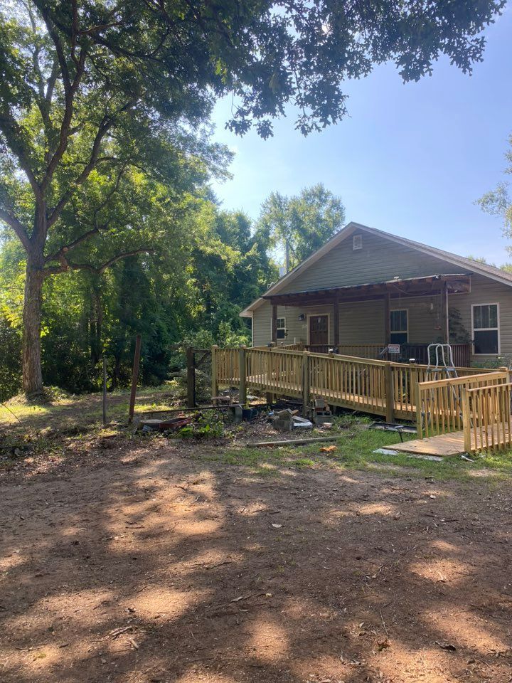 A single-story house with a wooden ramp for accessibility, set in a wooded area with trees and sunlight.