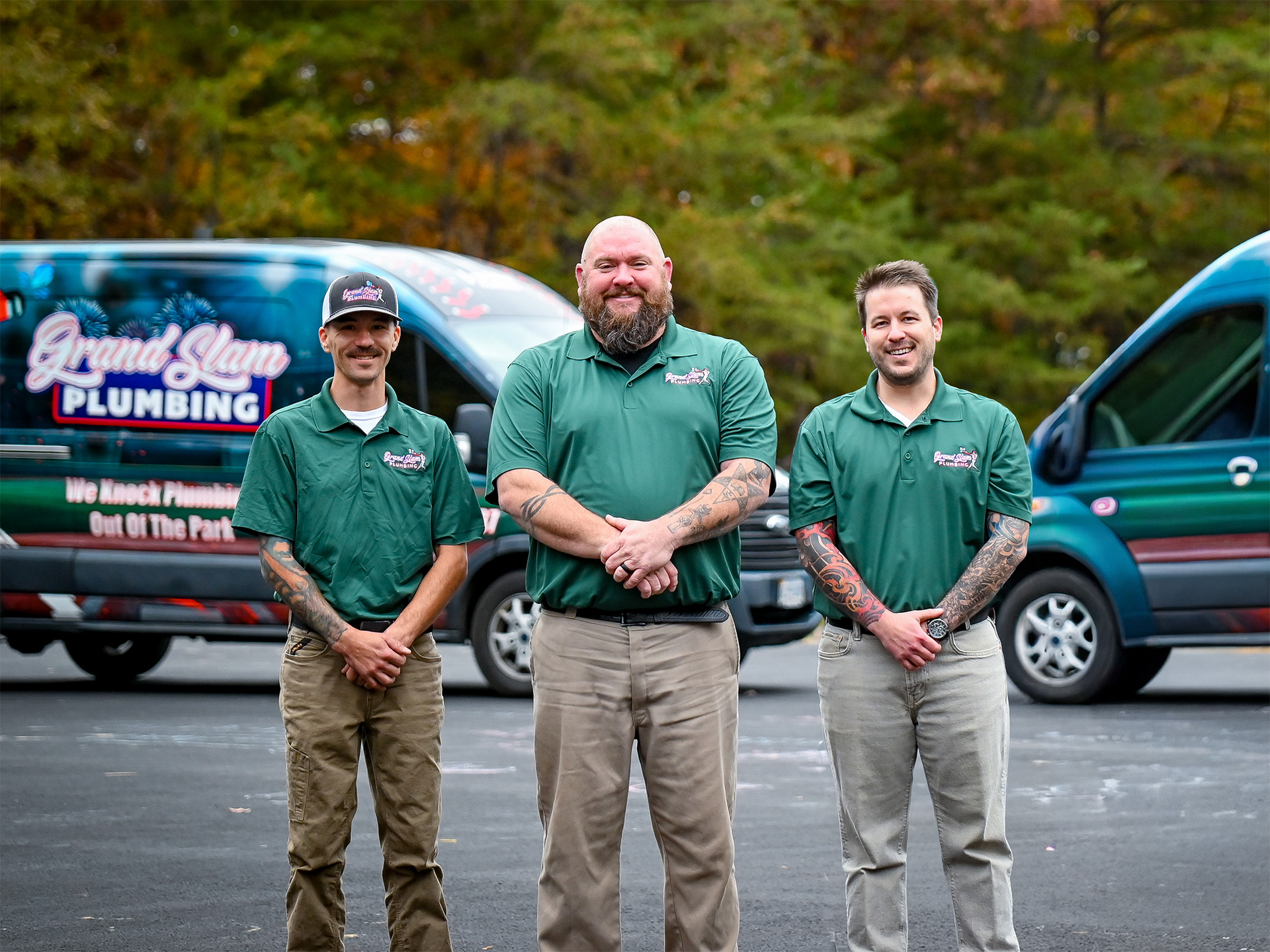 Three plumbers in green uniforms stand in front of branded vans on a parking lot.