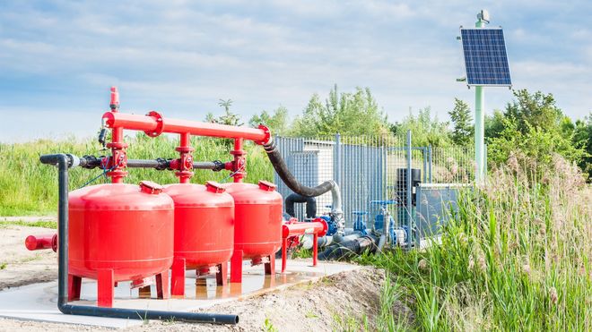 Red fire suppression tanks and piping beside a solar-powered pump in a grassy utility area.