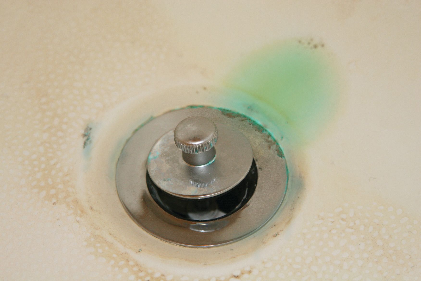 Close-up of a metal drain stopper with green residue around it in a white sink basin.