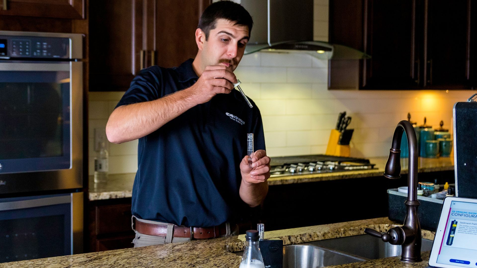 Man stirring a drink in a kitchen beside a stove and countertop electronics.